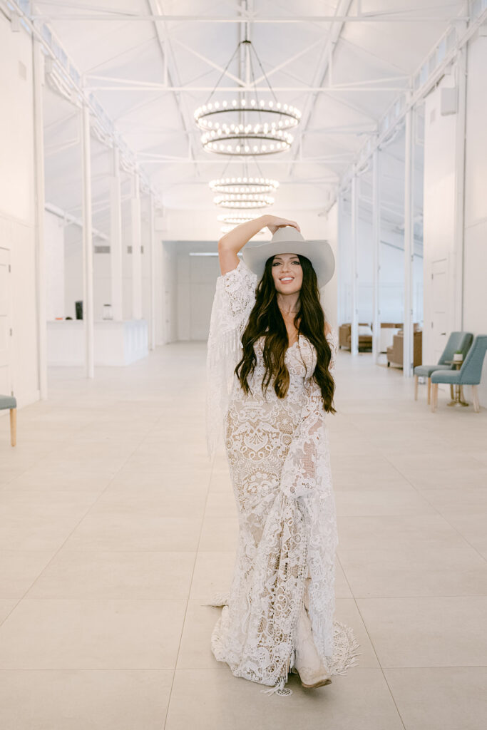 Bride in lace dress and hat smiling beneath chandelier inside The Videre’s renovated stables