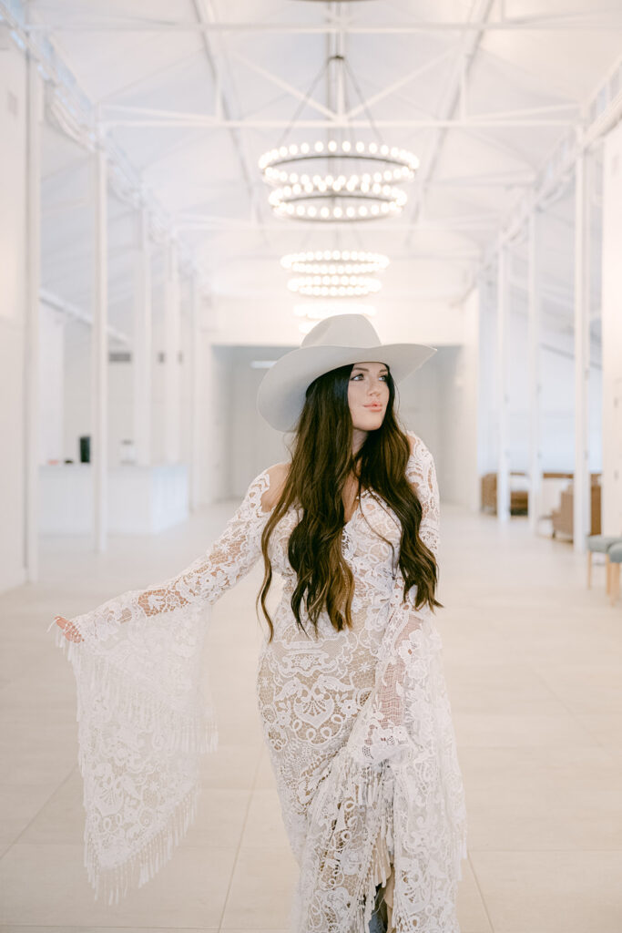 Bride in fringe lace gown posing confidently in chandelier-lit corridor at The Videre’s stables