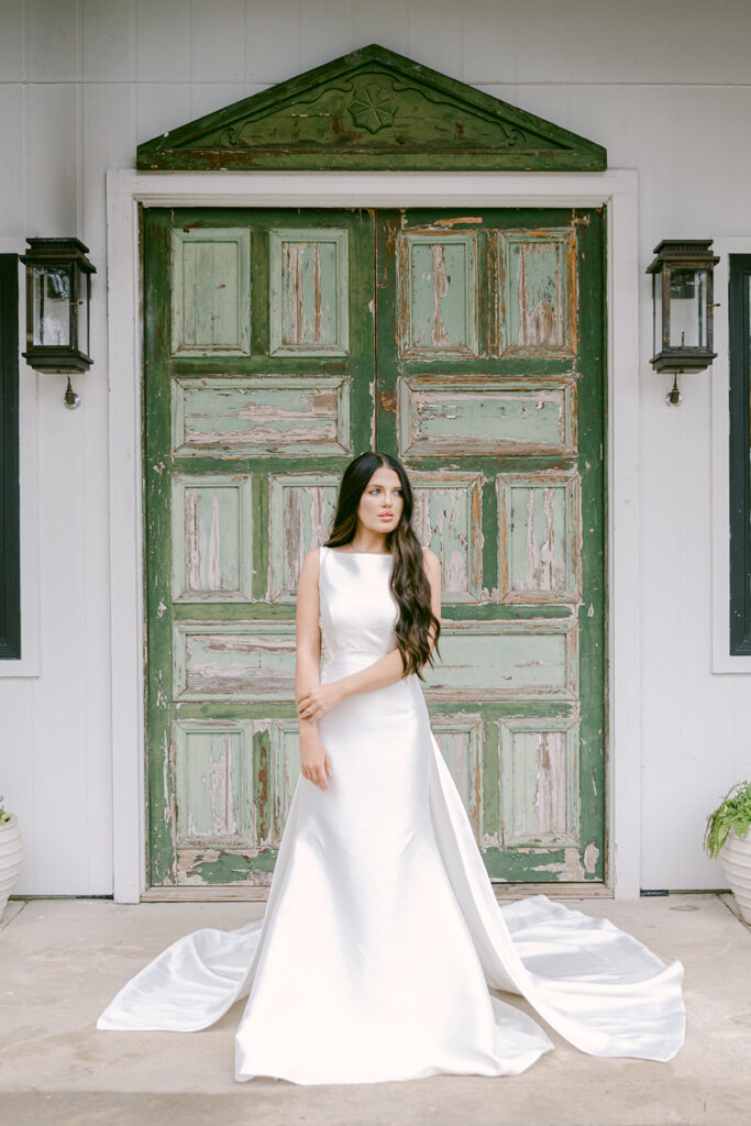 Bride standing with hands crossed in front of distressed green doors at The Château at The Videre wedding venue