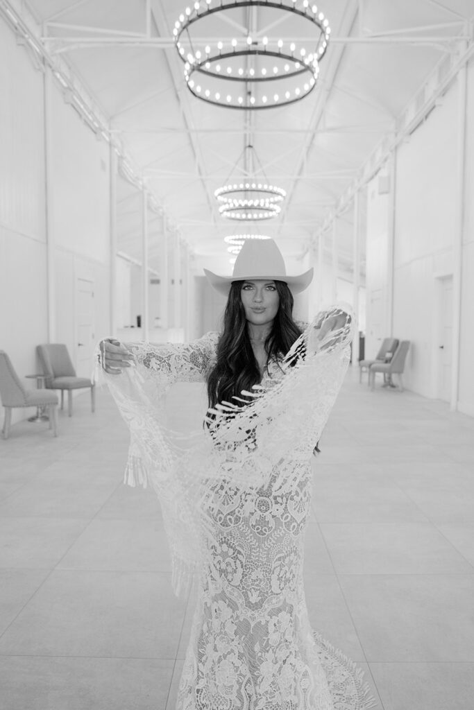 Black and white portrait of bride lifting fringe sleeves beneath chandelier lights at The Videre’s barn