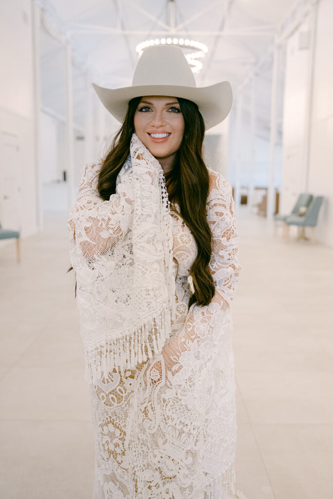 Close-up of bride smiling in cowboy hat and lace gown inside The Videre’s stables