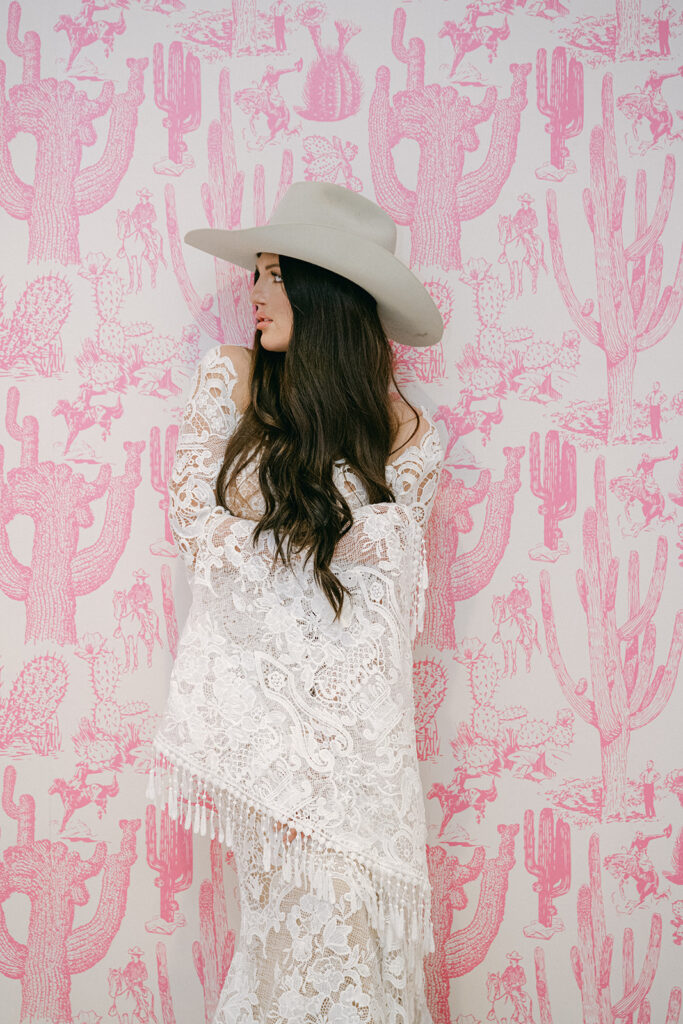 Bride in profile at pink cactus wall inside The Videre’s stables wearing fringe lace gown and hat
