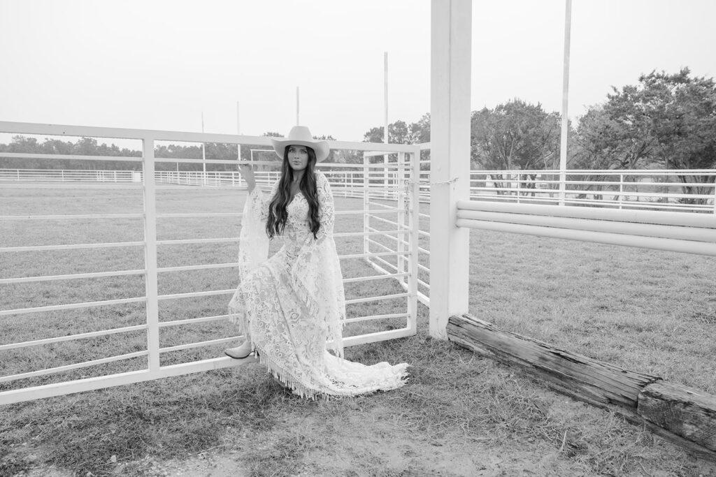 Bride leaning against white metal gate at The Videre’s open-air wedding arena in black and white
