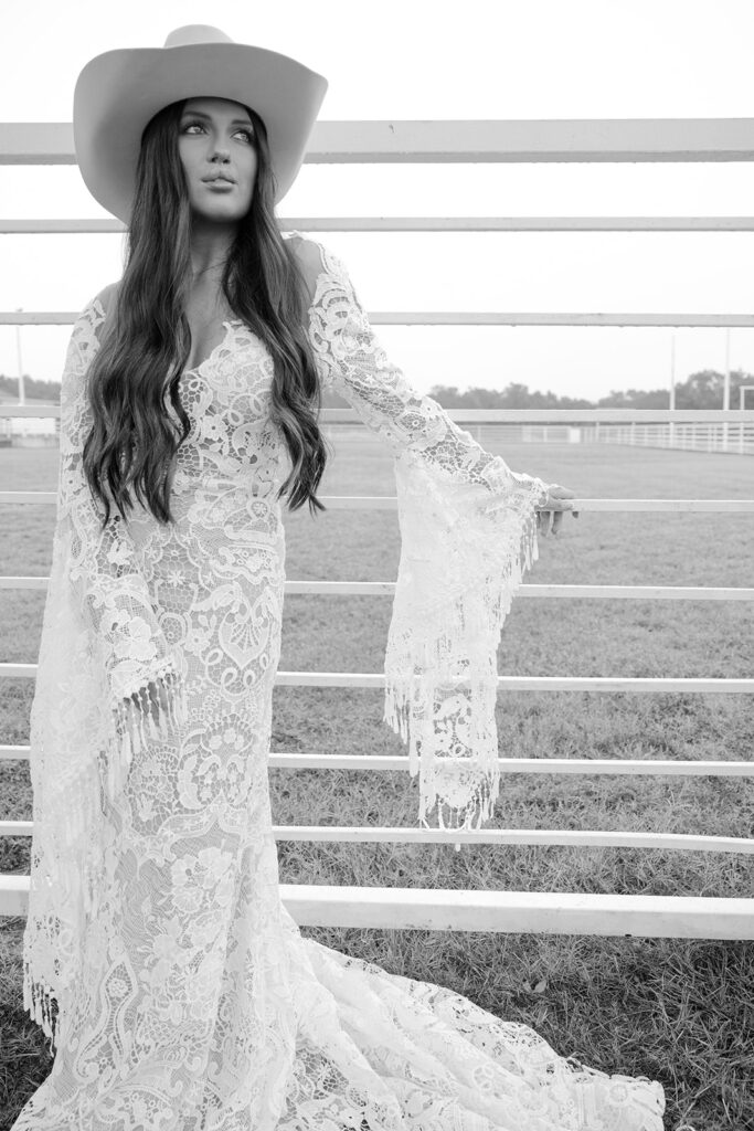 Black and white portrait of bride in western hat and lace dress near ranch fencing at The Videre