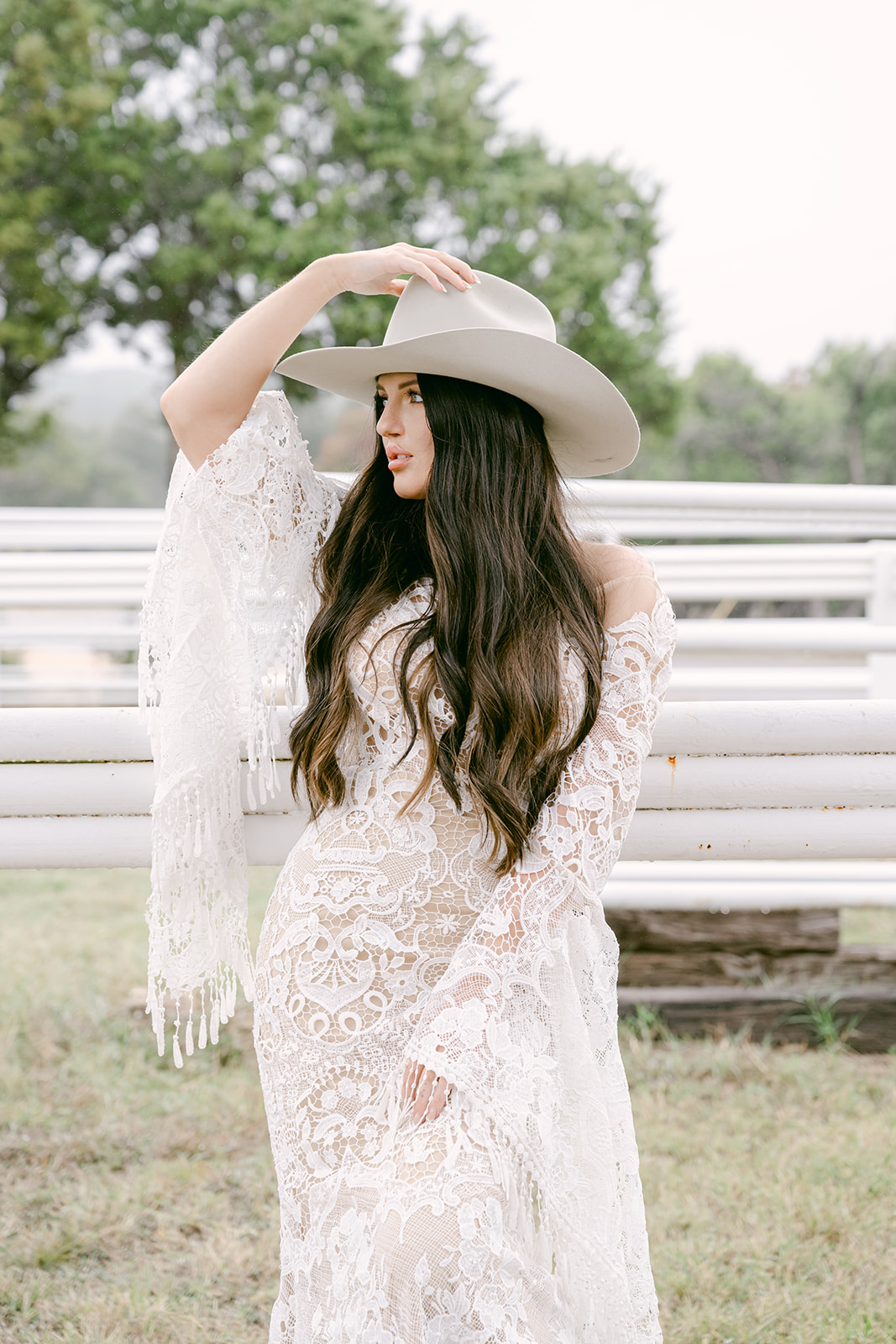 Close-up of bride wearing wide-brim hat and lace gown leaning on white arena fencing at The Videre