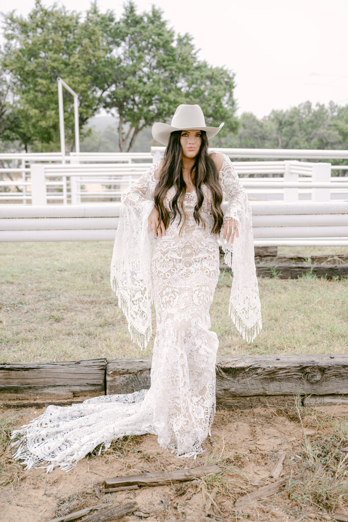 Bride in lace dress and wide-brim hat seated on log in The Videre’s open-air arena