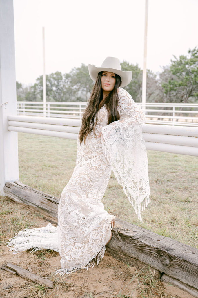 Bride leaning alongside ranch gate in fringe lace gown at The Videre’s arena