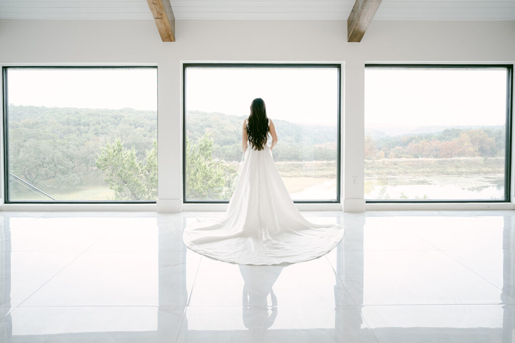 Bride standing inside The Videre wedding venue with long train and hill country views through large windows