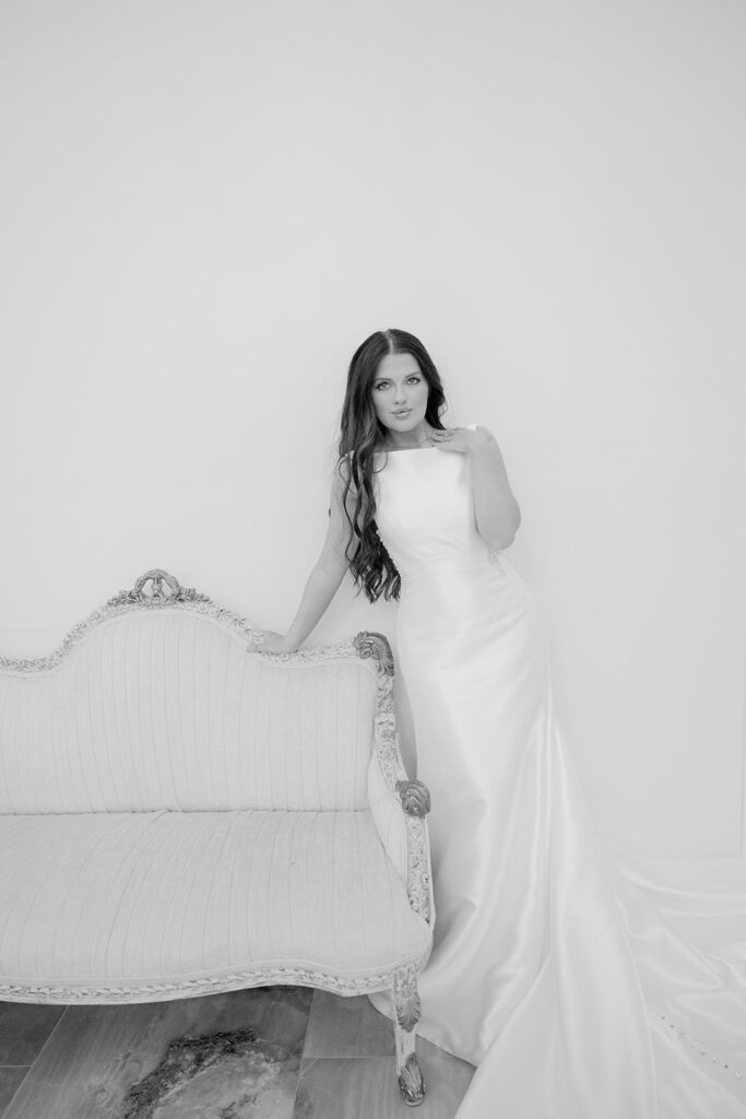 Bride in satin gown leaning against antique white couch in The Château bridal suite at The Videre