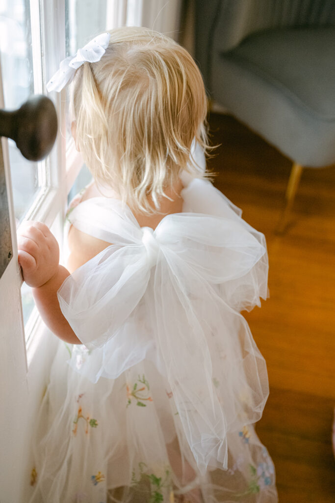 Toddler flower girl in white bow dress looking out the window at The Allen Farmhaus wedding venue
